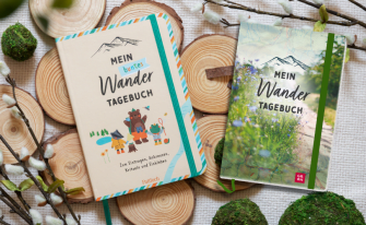 Ein dekoratives Flatlay auf einem strukturierten, weißen Stoff, umgeben von Holzscheiben und Weidenkätzchen-Zweigen. Es zeigt zwei verschiedene Designs eines Wandertagebuchs. Links ein cremefarbenes Buch mit dem Titel "MEIN buntes Wander TAGEBUCH" (Pattloch), das eine Gruppe von Cartoon-Waldtieren und eine Berggrafik zeigt. Rechts ein Buch mit dem Titel "MEIN Wander TAGEBUCH" (GROH), mit einem Coverbild einer Wiese voller Wildblumen und einer Berggrafik.