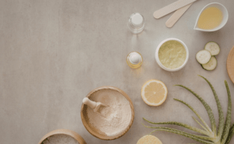 Draufsicht auf verschiedene natürliche Wellness- und Kosmetikzutaten, die auf einer grauen Steinfläche arrangiert sind. Zu sehen sind Holzschalen mit Pulver und Badesalz, eine Aloe-Vera-Pflanze, eine Zitronenscheibe, Gurkenscheiben, Öle in Fläschchen sowie Cremes und Holzspatel. Die linke Bildhälfte bietet freien Raum für Text.