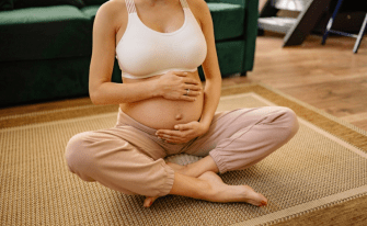 Eine schwangere Frau sitzt im Schneidersitz auf einer Matte auf dem Boden und hält sanft mit beiden Händen ihren unbedeckten Babybauch. Sie trägt ein weißes Sport-Top und eine bequeme, hellrosa Jogginghose. Der Kopf ist nicht im Bild zu sehen; im Hintergrund befindet sich ein grünes Sofa.