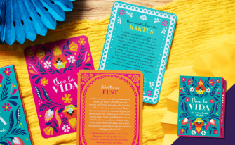 Bunter Hintergrund mit Papierblumen und drei Orakelkarten des Kartensets, im Vordergrund eine blaue rechte Ecke mit dem Cover der Box drauf