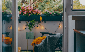 Gemütlicher Balkon bei Dämmerung mit leuchtender Lichterkette, Blumenkästen an der Brüstung, einer Topfpflanze und einem Klappstuhl mit senfgelbem Strickpullover und grüner Decke.