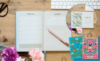 Aufgeschlagener Terminplaner 2027 auf Holzschreibtisch neben zwei Notizbüchern mit bunten Blumenstickereien.