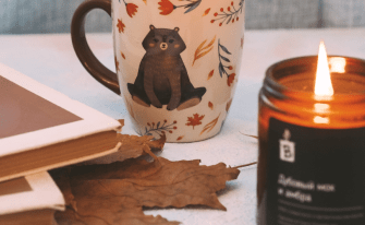 Gemütliches Stillleben mit einer brennenden Kerze im braunen Glas, einer Kaffeetasse mit Bärenmotiv und Blättern, getrockneten Herbstblättern und Stapeln von Büchern.
