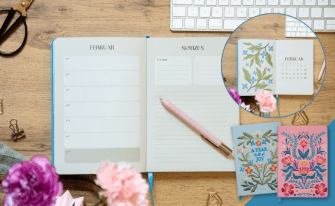 Aufgeschlagener Terminplaner auf Holzschreibtisch neben zwei Notizbüchern mit bunten Blumenstickereien.