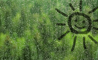 Regentropfen auf einer Glasscheibe, auf die eine lächelnde Sonne gezeichnet ist, mit unscharfem, grünem Hintergrund.