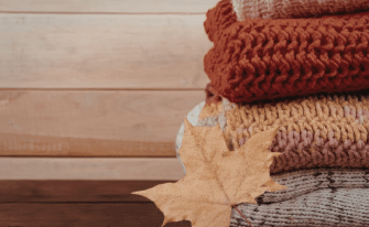Stapel aus dick gestrickten Pullovern in Herbstfarben (Rostrot, Gelb-Rosa und Grau) auf einer Holzoberfläche, davor liegt ein trockenes, braunes Ahornblatt.