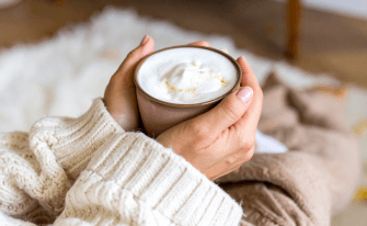 Nahaufnahme von zwei Händen im hellen Strickpullover, die eine warme Tasse Getränk mit einer dicken Milchschaumhaube und etwas Zimt- oder Gewürzpulver umfassen.