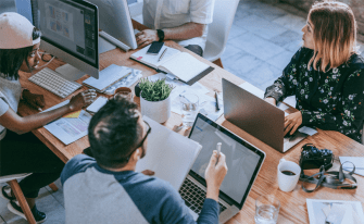 Mehrere Personen sitzen mit Laptop und anderen Büroartikeln an Tisch und diskutieren