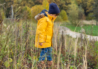 Ein kleiner Junge mit gelbem Parka, blauer Mütze, Jeans und Stiefeln geht über eine Wiese