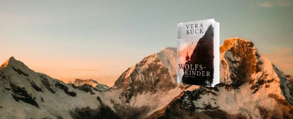 Das Buch-Cover zu „Wolfskinder“ von Vera Buck schwebt vor einer gewaltigen, schneebedeckten Bergkette, die vom warmen, sanften Licht der Abendsonne angestrahlt wird.