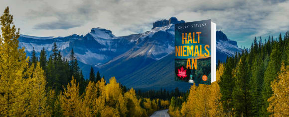 Das Buch-Cover zu „Halt niemals an“ von Chevy Stevens schwebt über einer einsamen Straße, die durch einen herbstlich gefärbten Nadelwald führt, während sich im Hintergrund eine gewaltige, schneebedeckte Bergkette unter blauem Himmel erstreckt.