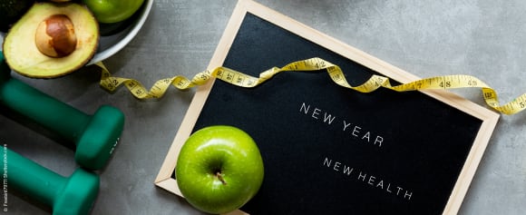 Ein Schild mit der englischen Aufschrift Neues Jahr, neue Gesundheit. Neben dem Schild liegen Hanteln, ein Maßband, ein Apfel und eine Avocado. 