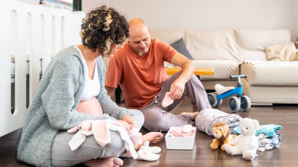 Schwangere Frau und Mann sitzen auf dem Boden neben einem Babybett und sortieren Babykleidung