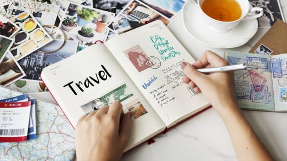 Das Bild zeigt ein aufgeschlagenes Blanko-Buch, in welches eine Frau gerade Erinnerungen an eine Reise niederschreibt. Drumherum liegen viele Fotos und eine Tasse Tee steht oben rechts im Anschnitt. Die Frau hält einen Stift in der Hand.