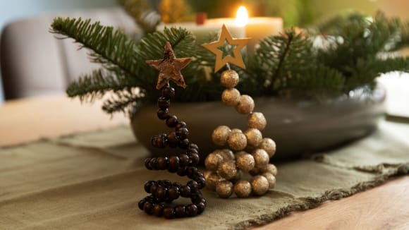 Zwei gebastelte Tannenbäume aus Holzperlen auf einem Holztisch, ein Tischläufer und ein weihnachtliches Gesteck sorgen für das passende Ambiente 