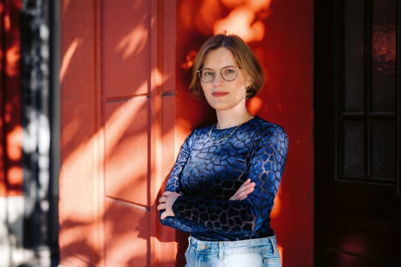 Friederike Oertel steht lächelnd und mit verschränkten Armen draußen vor einer orangenen Wand