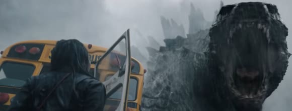 Monarch: Legacy of Monsters-Screenshot. Links sehen wir einen Mensch mit halblangen Haaren von Hinten vor einem Schulbus, in der rechten Bildhälfte guckt Godzilla mit aufgerissenem Maul in die Kamera