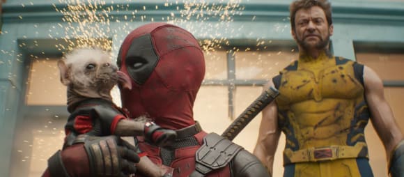 Im Vordergrund Deadpool in seinem anzug, der von einem kleinen, haarlosen Hund mit langer Zunge im Gesischt abgeschlabbet wird. Im Hintegrund Wolverine in gelbem Dress, der eine grimmige Fratze schneidet. Über allem fliegen kleine Funken.