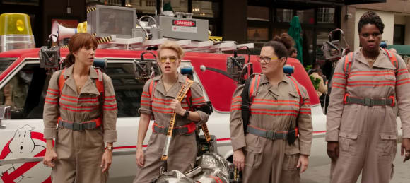 Die vier weiblichen Ghosbusters aus dem Film von 2014 in voller Montur vor dem Ghostbusters-Mobil stehend
