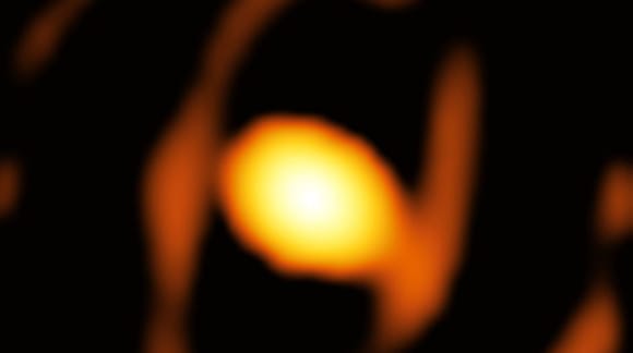 Verschwommene Aufnahme eines Sterns kurz vor der Explosion zur Supernova. In der Mitte ein heller, gelber Kreis, umgeben von einem gelblichen Schatten in Form einer Acht, dazwischen schwarzer Hintergrund.