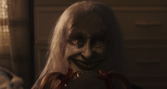 Dämonische alte Frau mit langen weißen Haaren blickt böse grinsend in die Kamera, vor sich hält sie eine Spielzeugpuppe, von der nur ein Teil des Kopfes zu sehen ist.