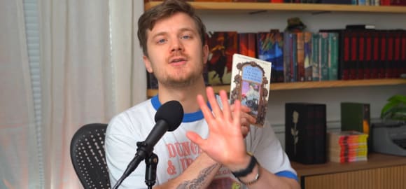 Ein junger Mann in einem Dungeons-and-Dragons-T-Shirt sitzt blickt zu uns (also der Kamera) hinter sich ein Bücherregal, vor sich ein Mikrofon, in der linken Hand hält er ein kleines Taschenbuch, die rechte Hand ist davor ausgestreckt.