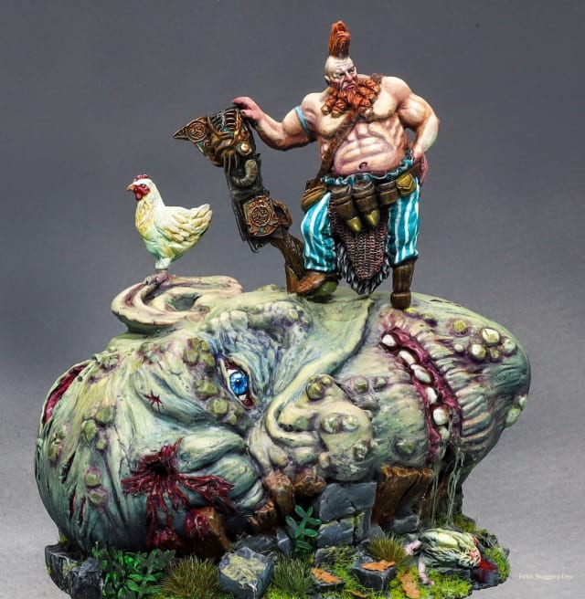 Fantasyminiatur - Keltischer Krieger steht mit nacktem, muskulösem Oberkörper in blauer Hose auf einem Felsen. Rechts von ihm ein Huhn.