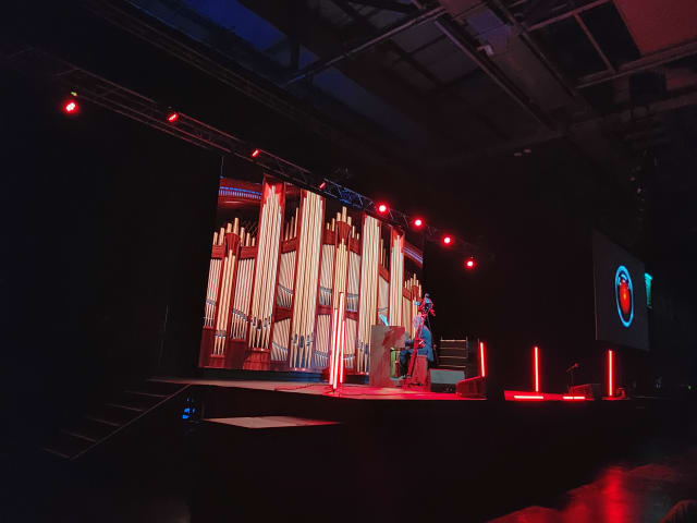 Ein Musiker mit grauen Haaren sitzt auf der Bühen an einer Orgel mit hohen rötlich beleuchteten Orgelpfeifen.