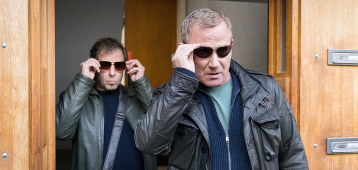 Ekki Talkötter (Oliver Korittke) und Overbeck (Roland Jankowsky) kommen gemeinsam aus einem Hauseingang. Beide tragen Lederjacken und setzen sich im Gehen gerade eine dunkle Sonnenbrille auf.