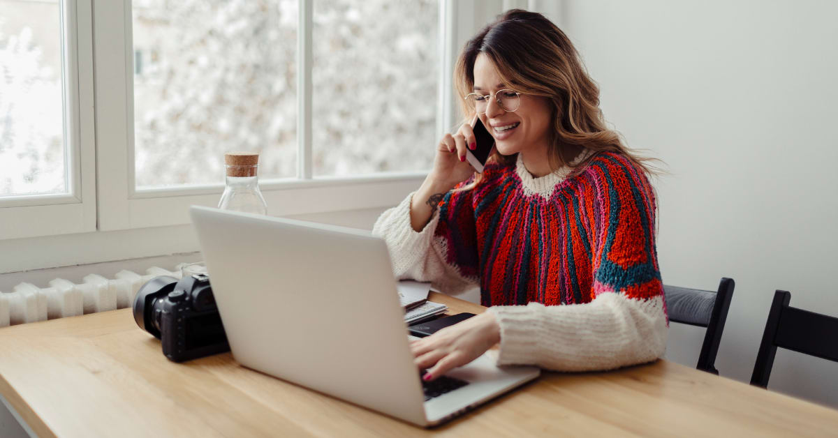 A white woman sitting at a table is looking down at a laptop and smiling while on the phone
