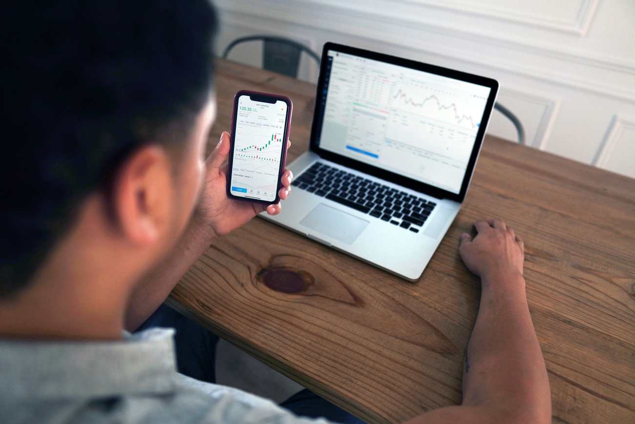Photo shows the back of a man's head as he looks at a phone and laptop showing his freelance finances.