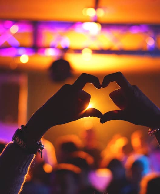 Person holding up a heart with their hands in front of a blurry concert