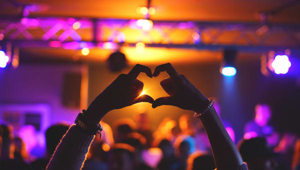 Person holding up a heart with their hands in front of a blurry concert