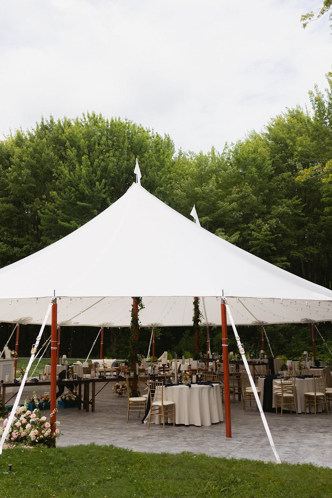 White sailcloth wedding tent sheltering reception tables with gold chiavari chairs and floral arrangements, surrounded by gre