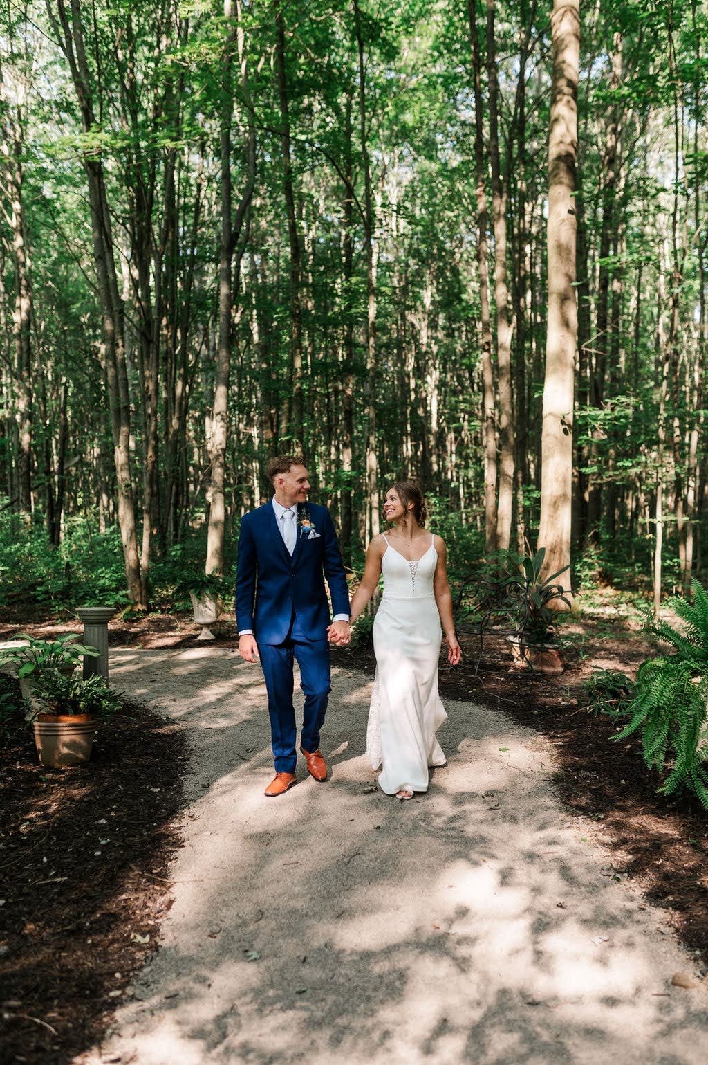 Bride and groom walking hand-in-hand along a sunlit wooded garden path at Sugar Lake Weddings
