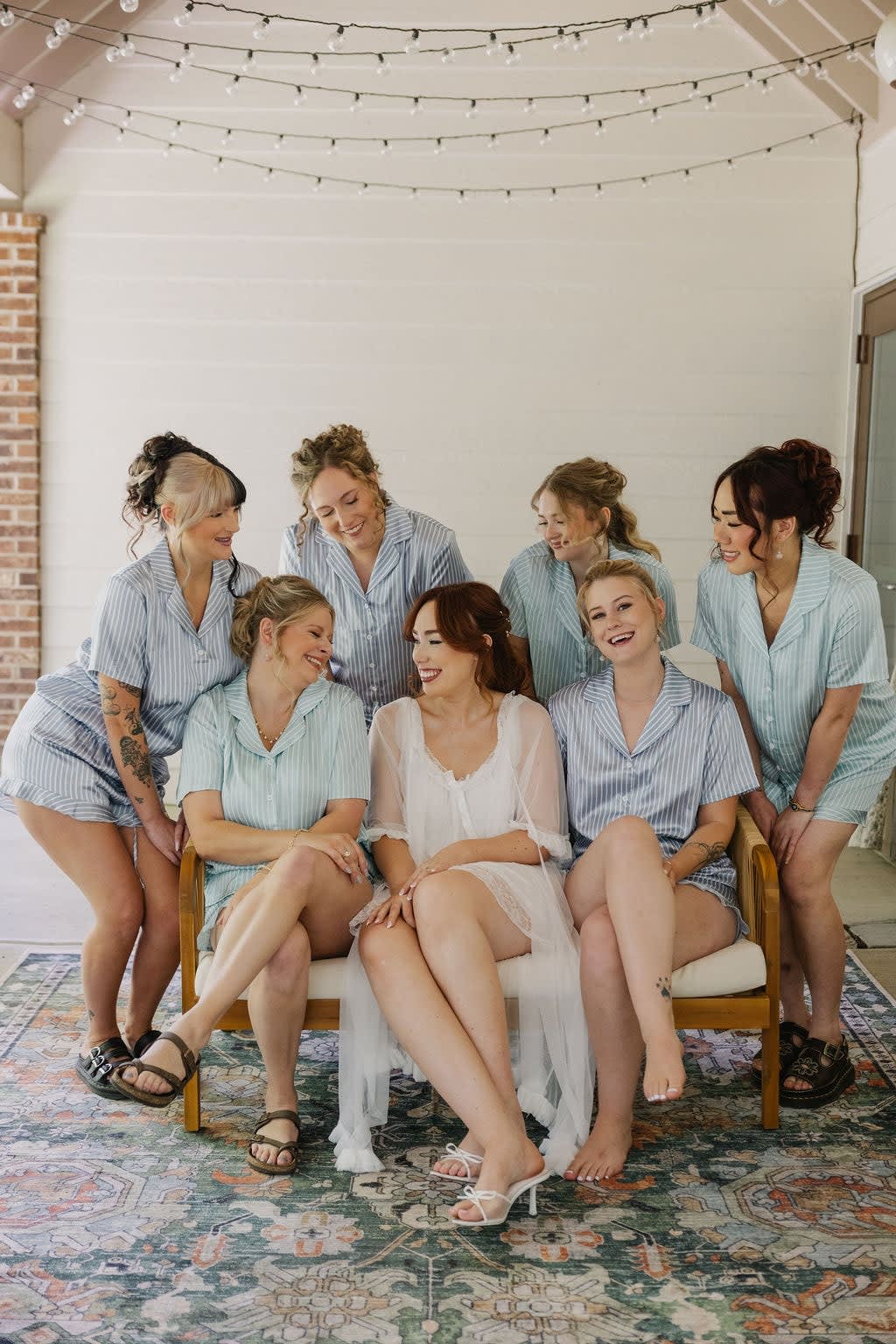 Bride in white robe seated with bridesmaids in striped pajamas laughing under string lights at Sugar Lake Weddings
