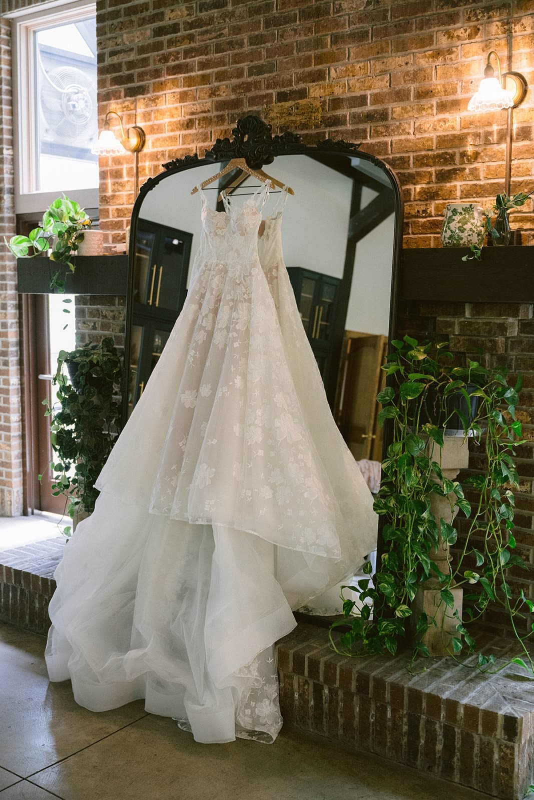Floral appliqué wedding gown hanging from an ornate black mirror above a brick fireplace flanked by trailing greenery