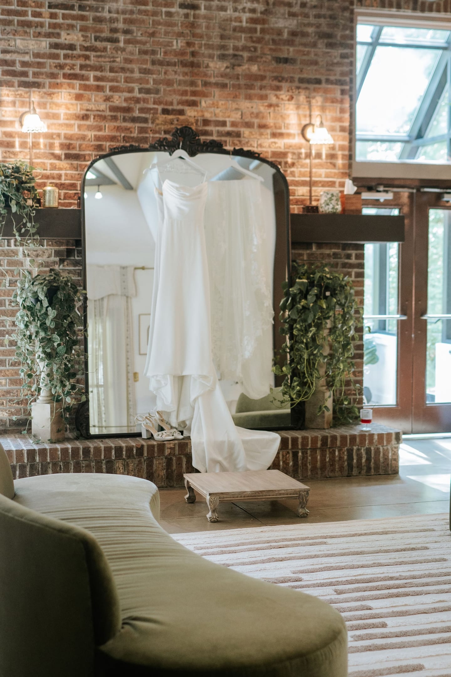 White wedding dress hanging on an ornate black mirror in a rustic brick bridal suite with green velvet furniture