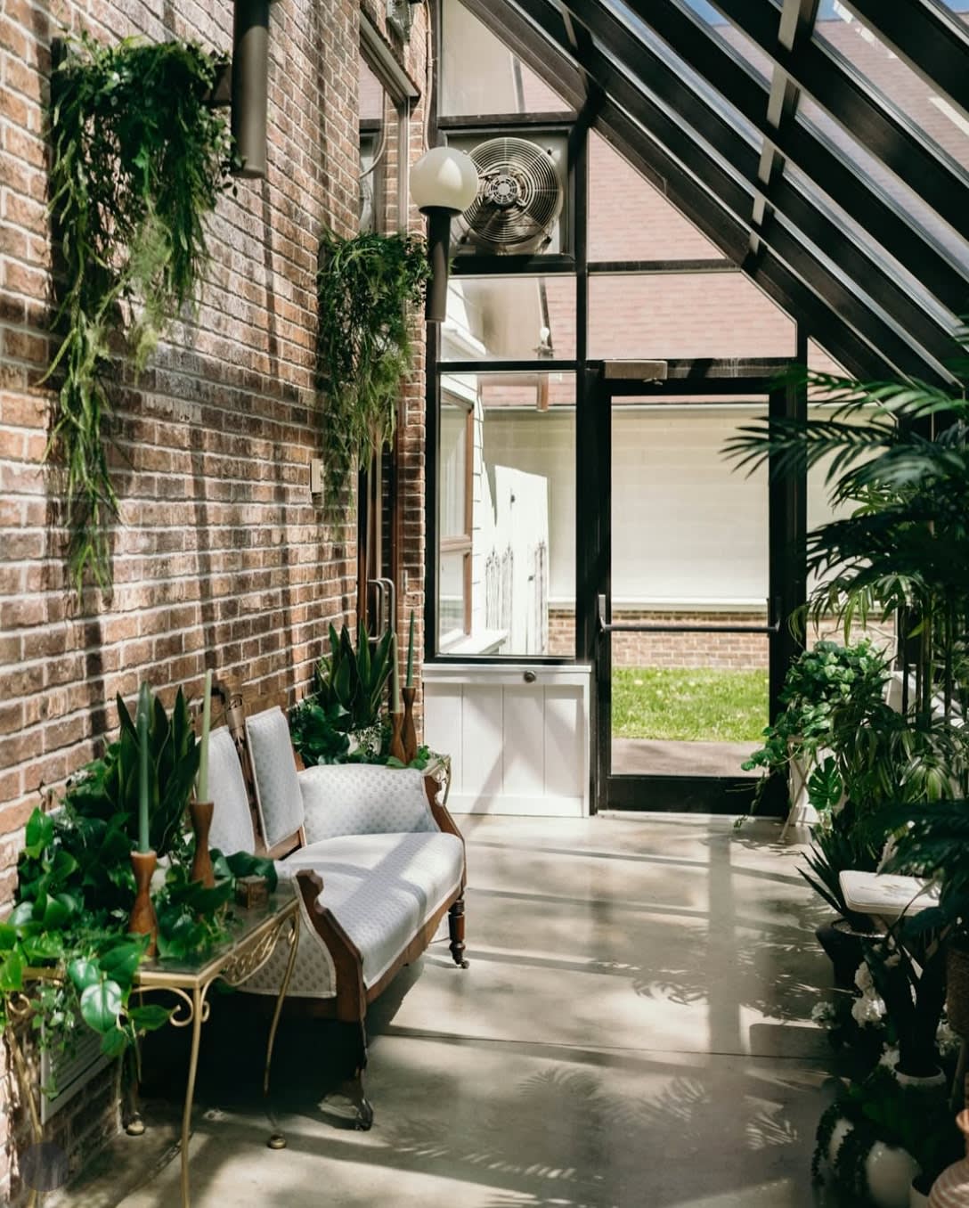 Sunlit conservatory with exposed brick wall, lush greenery, and an antique white settee at Sugar Lake Weddings