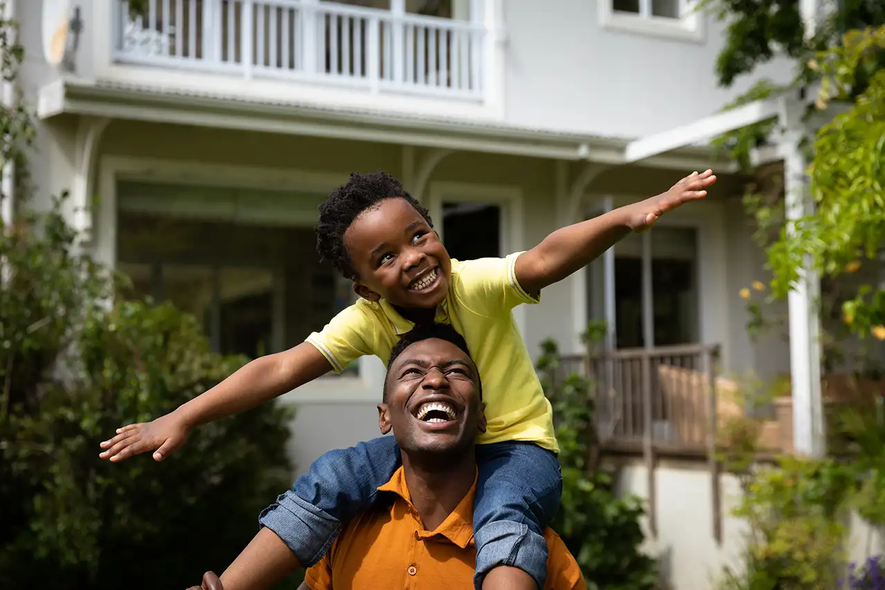 Young boy with outstretched arms flying on shoulders of dad in front of home