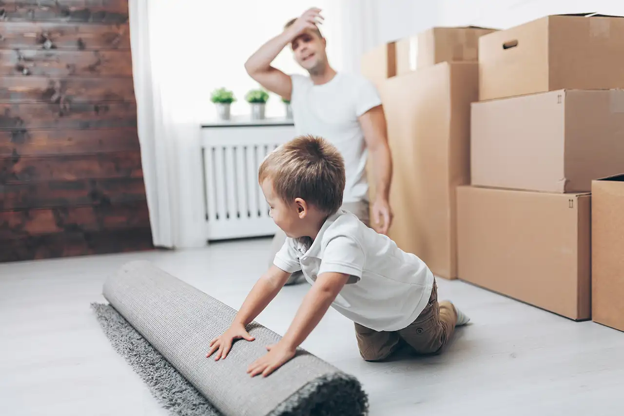 Father watching toddler son roll up rug in front of packing boxes