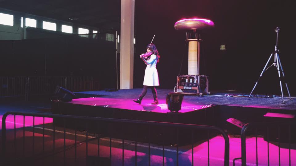 Kaitlyn Hova playing violin on stage next to a tesla coil