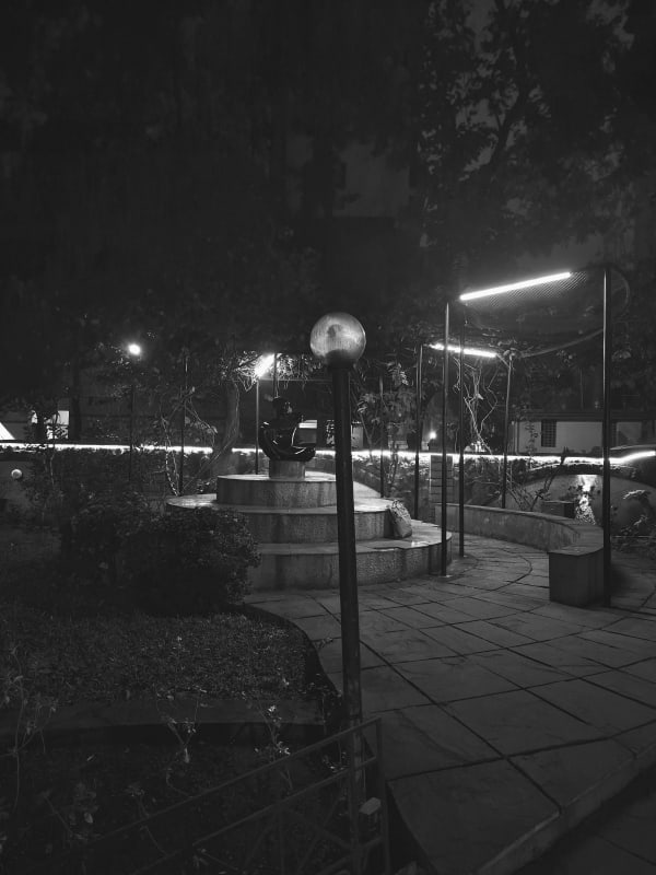 A landscaped garden at night with a glowing globe lamp, a seated sculpture on a stepped platform and a lit pergola, in black and white.