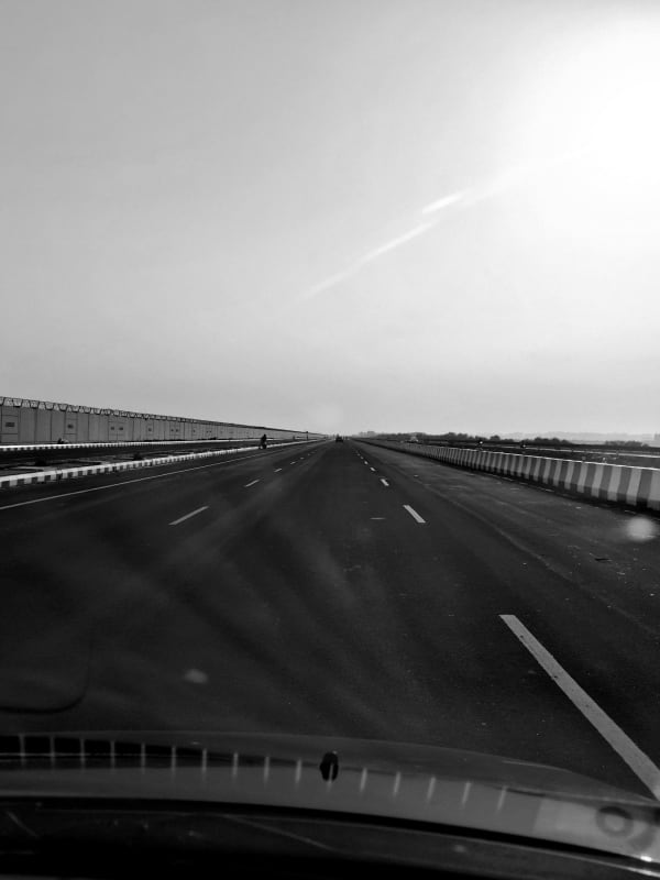 An empty multi-lane expressway stretching to the horizon under a bright sky, seen through a car windshield, in black and white.
