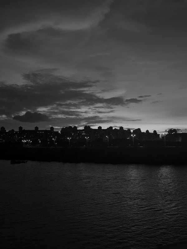 A city skyline in silhouette across open water at dusk under a dramatic cloudy sky, streetlights glowing along the far bank, in black and white.