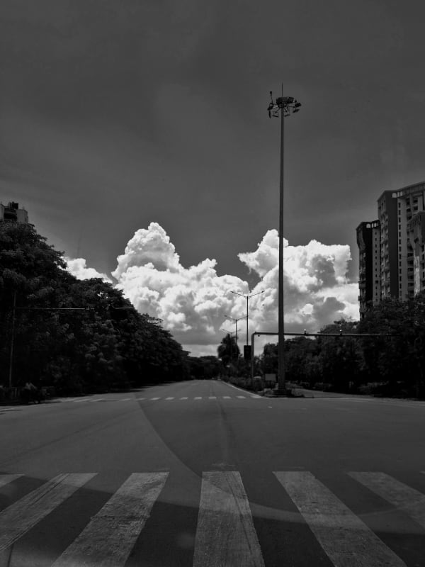 An empty road with a zebra crossing in the foreground, a tall lamppost and towering cumulus clouds filling the sky, in black and white.
