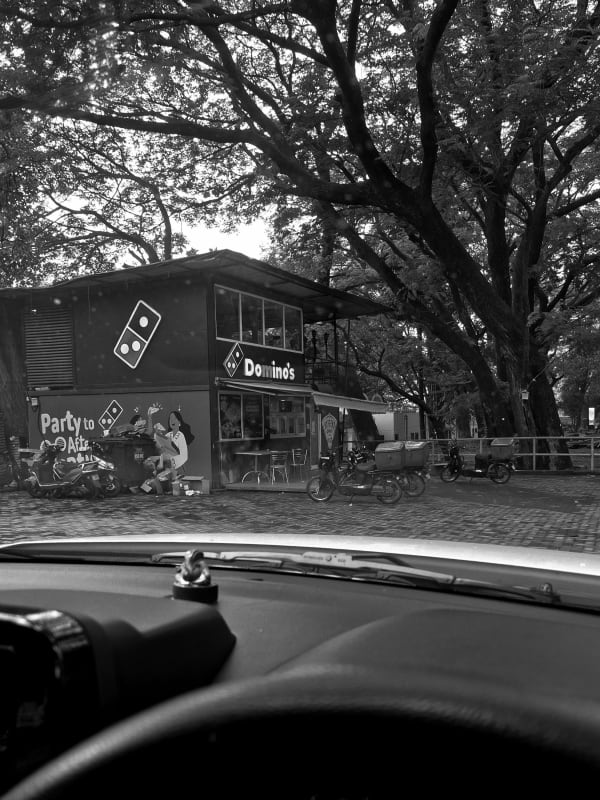 A Domino's Pizza outlet beneath large spreading trees with delivery bikes parked outside, seen from inside a car, in black and white.
