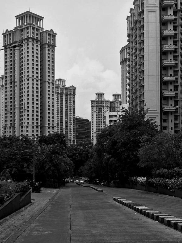 An empty tree-lined road running between tall residential high-rise towers under an overcast sky, in black and white.