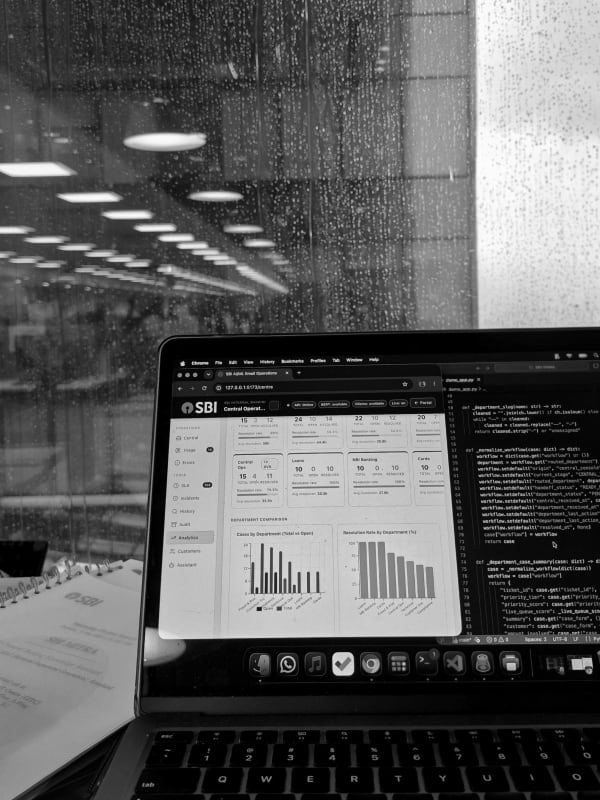 A laptop on a desk showing an operations dashboard beside a window of Python code, rain-streaked office glass behind it, in black and white.