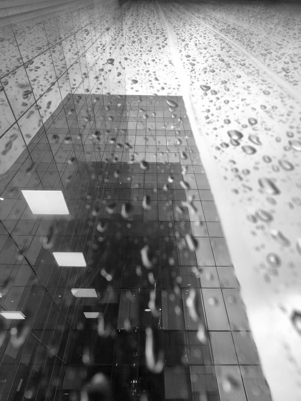 Raindrops on a glass pane with the reflection of a tiled high-rise facade and ceiling lights beyond, an abstract study in black and white.